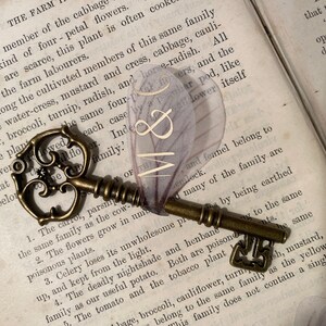 May include: A vintage skeleton key with a decorative floral design, resting on an open book with text about different types of plants and flowers. The key is made of brass and has a worn, antique finish. The book is open to a page with the text "THE FARM" at the top.