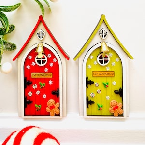 May include: Two decorative wooden elf workshop doors. One is red, the other yellow. Both have a white frame, a small bell, and the text "ELF WORKSHOP". Each door is decorated with snowflakes, holly, and a gingerbread man.
