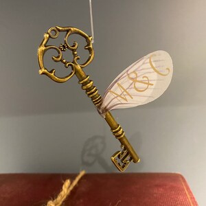 May include: A gold-toned key with a decorative design and a white, translucent fairy wing attached. The wing has the initials "M & C" written on it.