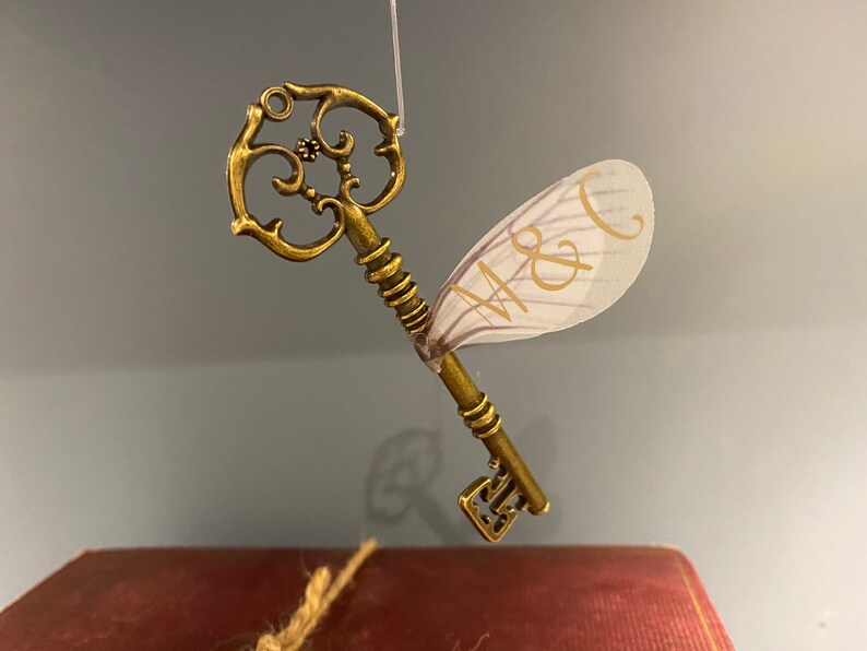 May include: A gold-toned key with a decorative design and a white, translucent fairy wing attached. The wing has the initials "M & C" written on it.