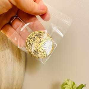 Puede incluir: Una moneda navide&ntilde;a dorada con el texto "I Met Santa" en una bolsa de pl&aacute;stico transparente. La moneda presenta una ilustraci&oacute;n de Pap&aacute; Noel y elementos decorativos. La moneda es redonda y tiene un borde detallado.