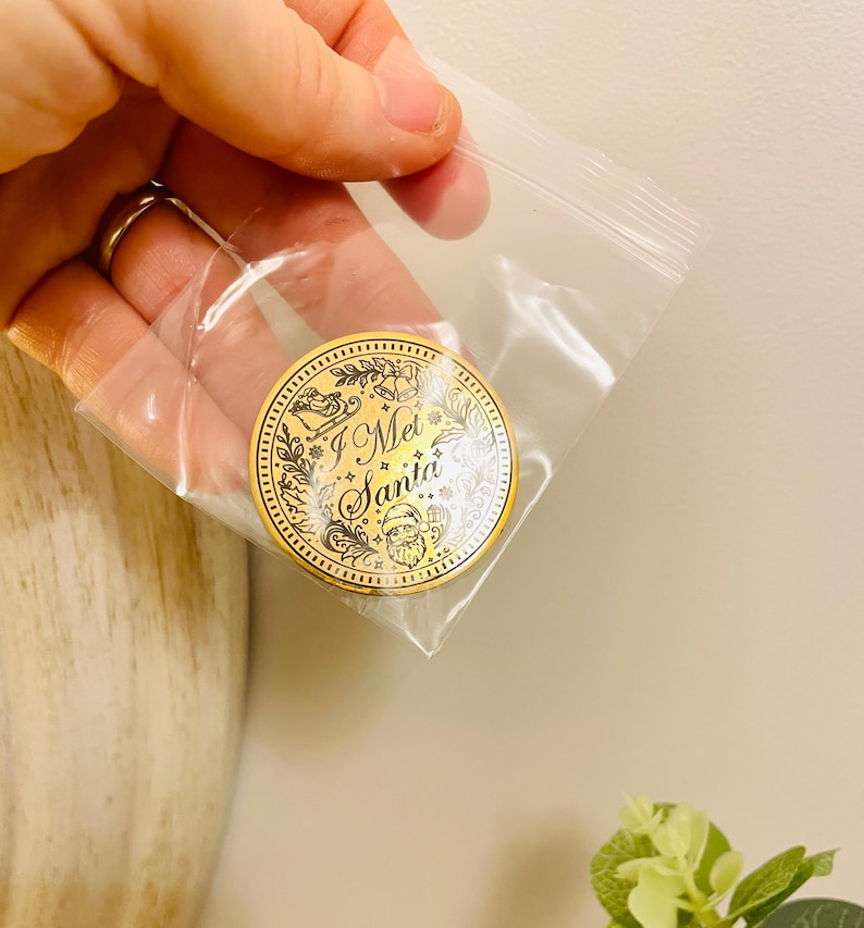 Puede incluir: Una moneda navide&ntilde;a dorada con el texto "I Met Santa" en una bolsa de pl&aacute;stico transparente. La moneda presenta una ilustraci&oacute;n de Pap&aacute; Noel y elementos decorativos. La moneda es redonda y tiene un borde detallado.