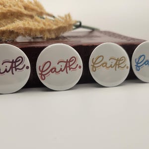 May include: Four white round buttons with the word "Faith" in a cursive font. Each button has a different color for the word: burgundy, red, gold, and blue. The buttons are on a dark brown surface.