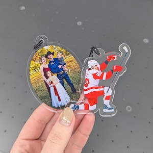 Puede incluir: Adorno acr&iacute;lico transparente con una foto familiar ovalada y un jugador de hockey con uniforme rojo y blanco. El jugador de hockey est&aacute; arrodillado con un palo.