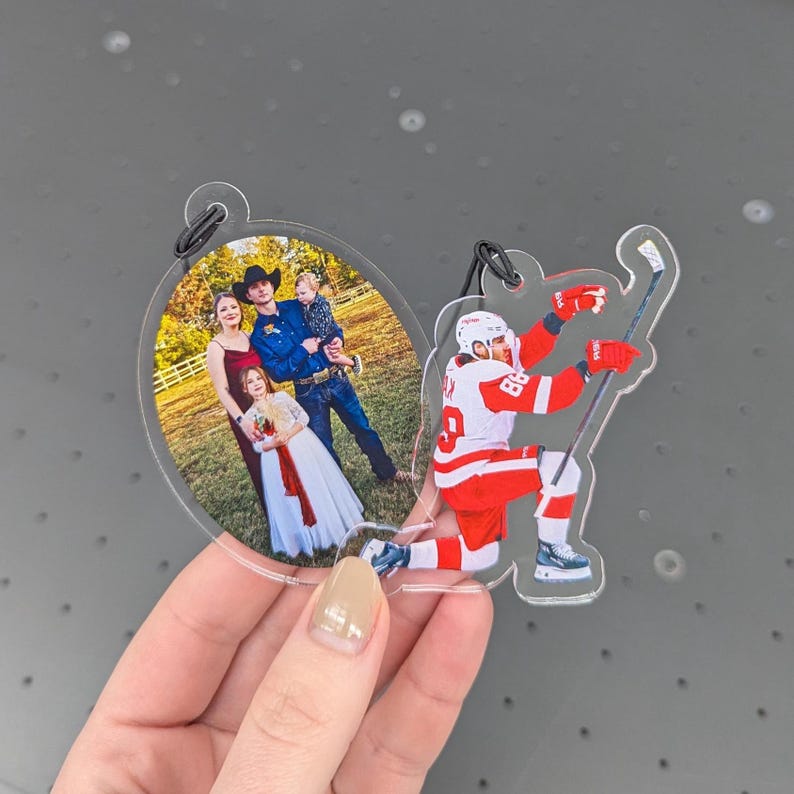 Puede incluir: Adorno acr&iacute;lico transparente con una foto familiar ovalada y un jugador de hockey con uniforme rojo y blanco. El jugador de hockey est&aacute; arrodillado con un palo.
