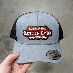 May include: A heather gray and black mesh trucker hat featuring a central patch with the text Grosenbaugh Farms KETTLE CORN Louisville, Ohio and a small kettle graphic. A circular black and silver sticker is on the brim reading The Authentic Yupoong Snapback.