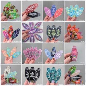 May include: A collection of sixteen sets of motel-style keychains featuring various colors, glitter finishes, and printed text. Each keychain has a distinct elongated shape with a hole at the top, displaying phrases like Cozy Brother, The Wax Coven Club, and Emma.