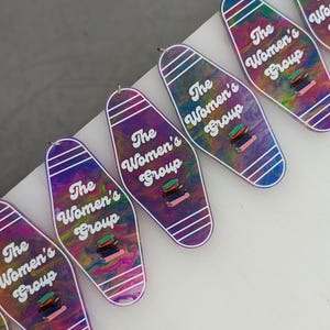 May include: Multiple motel-style keychains featuring a swirling purple, blue, and yellow marbled pattern. Each keychain displays the white text The Women's Group above a small graphic of a stack of colorful books, with white stripes at the ends.
