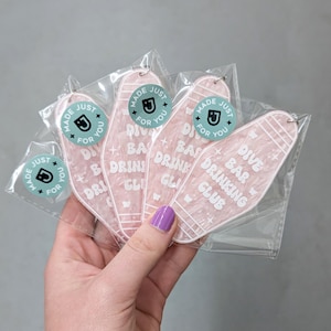 May include: Four motel-style keychains made of translucent pink marbled acrylic, each featuring the white text 'DIVE BAR DRINKING CLUB' and small star icons. Each keychain is individually packaged in a clear plastic sleeve with a round sticker.