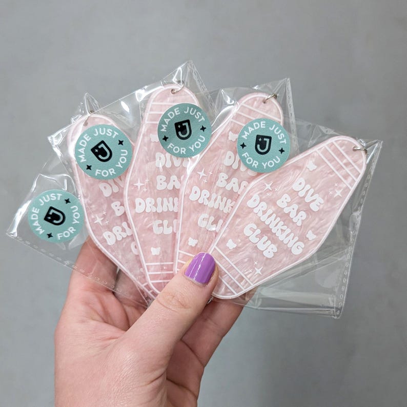 May include: Four motel-style keychains made of translucent pink marbled acrylic, each featuring the white text 'DIVE BAR DRINKING CLUB' and small star icons. Each keychain is individually packaged in a clear plastic sleeve with a round sticker.