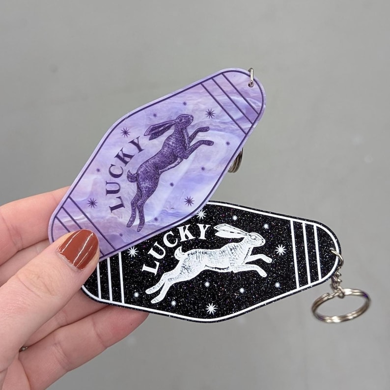 Op de afbeelding: Twee sleutelhangers in motelstijl met een afbeelding van een springend konijn en het woord LUCKY. De ene is paars met een gemarmerde afwerking, de andere is zwart met een glittertextuur. Beide hebben een puntige ovale vorm en een metalen ring.