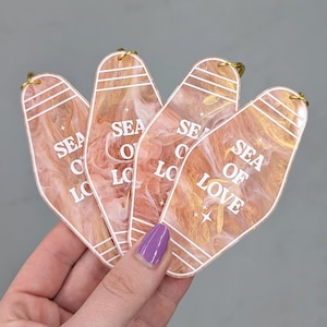 May include: Four motel-style keychains with a peach and gold marbled pattern. Each keychain features the white text "SEA OF LOVE" with small star accents and three horizontal white lines at the top. Each piece includes a gold metal jump ring.