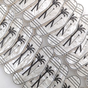 May include: A collection of elongated, oval-shaped white acrylic tags with a pearlescent finish. Each tag features a black graphic of a palm tree, horizontal lines, and the letter N. Each piece includes a small hole at the top for attachment.