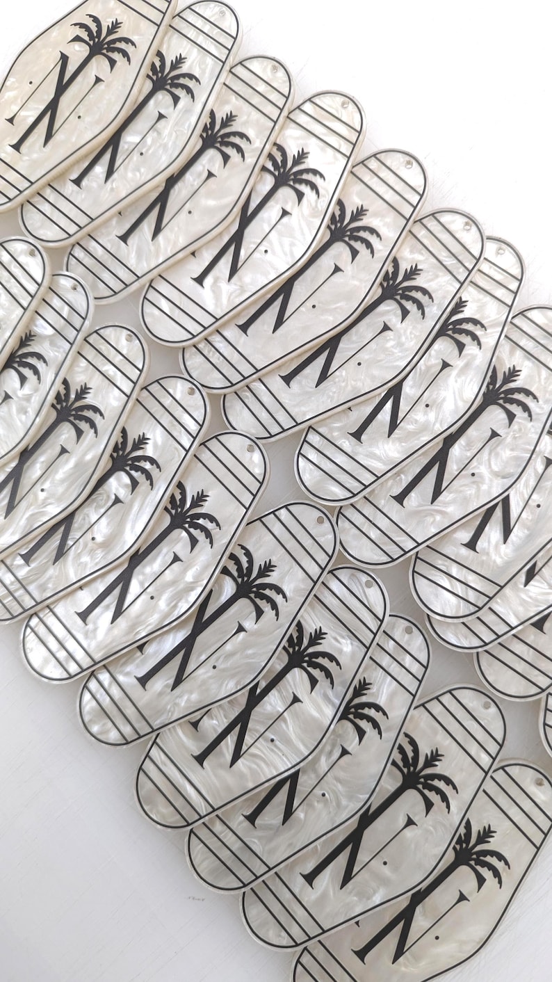 May include: A collection of elongated, oval-shaped white acrylic tags with a pearlescent finish. Each tag features a black graphic of a palm tree, horizontal lines, and the letter N. Each piece includes a small hole at the top for attachment.