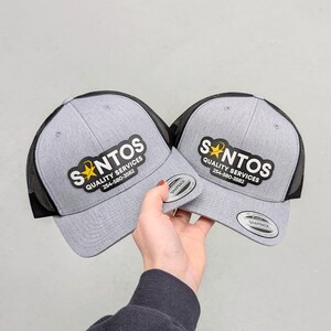 May include: Two gray heather snapback caps with black mesh backs. Each cap features a black patch with the white text SANTOS QUALITY SERVICES, a yellow star icon, and the phone number 254-580-3582. A circular sticker is on the brim of each cap.