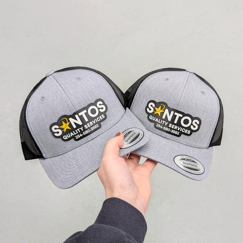 May include: Two gray heather snapback caps with black mesh backs. Each cap features a black patch with the white text SANTOS QUALITY SERVICES, a yellow star icon, and the phone number 254-580-3582. A circular sticker is on the brim of each cap.