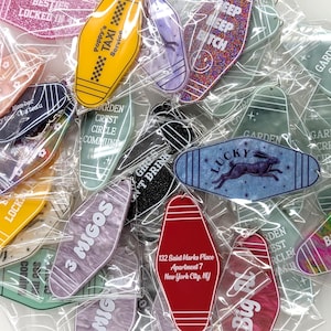 May include: A collection of assorted motel-style keychains in individual plastic sleeves, featuring various colors, patterns, and printed text including Poppy's Taxi Service, Lucky with a rabbit graphic, 3 Migos, and 132 Saint Marks Place Apartment 7.