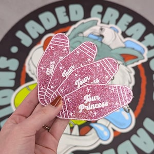 May include: Three pink glittery motel-style keychains shaped like elongated ovals with white stripes at the ends. Each keychain features the white text "Tour Princess" printed in a cursive font, accented by small white star shapes across the surface.