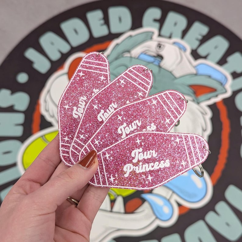 May include: Three pink glittery motel-style keychains shaped like elongated ovals with white stripes at the ends. Each keychain features the white text "Tour Princess" printed in a cursive font, accented by small white star shapes across the surface.