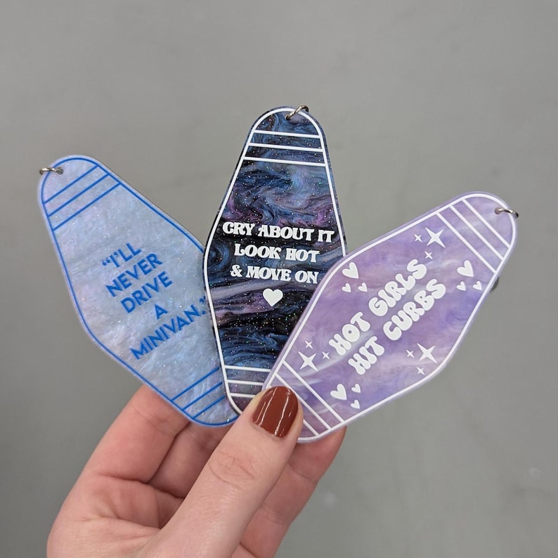 May include: Three motel-style keychains with rounded tops. The left is light blue with "I'LL NEVER DRIVE A MINIVAN." The center is dark blue and purple marble with "CRY ABOUT IT LOOK HOT & MOVE ON." The right is purple with "HOT GIRLS HIT CURBS."