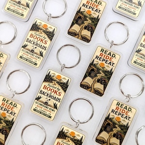 May include: Rectangular acrylic keychains featuring two distinct designs. One design shows an open book with the text 'FUELED BY BOOKS & BACKROADS SECONDHAND ROWS BOOK SHOPPE MT. CARMEL, PA 2026 EDITION'. The other shows a landscape with 'READ. RIDE. REPEAT.'