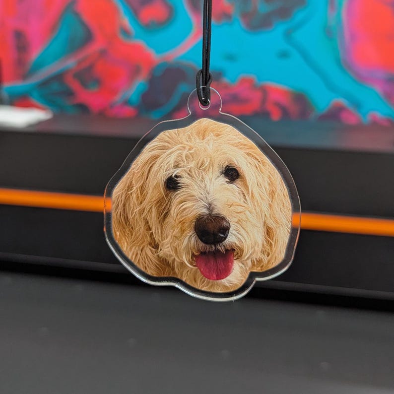 Puede incluir: Un adorno acr&iacute;lico transparente con un perro golden doodle sonriente con la lengua rosa. El perro tiene pelaje marr&oacute;n claro y ojos negros. El adorno cuelga de una cuerda negra.