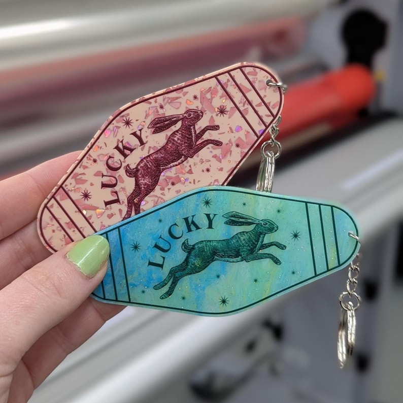 Op de afbeelding: Twee sleutelhangers in motelstijl met een afbeelding van een springend konijn en het woord LUCKY. De ene is lichtroze met een glinsterend, gefragmenteerd patroon, de andere is een blauw-groen verloop met een subtiele glans en metalen ringen.