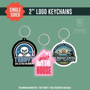 May include: Three single-sided 2-inch logo keychains. One is black with a white bear logo and text "TEDDY'S JIU JITSU ACADEMY". Another is pink, house-shaped, with "girl! BUY THE HOUSE" text. The third is white with a bridge logo and text "THE BRIDGE CENTER".