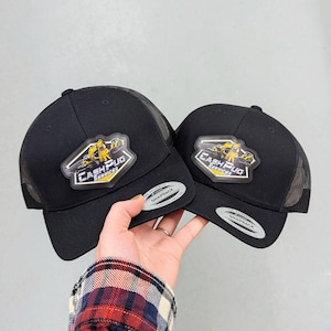 May include: Two black snapback trucker hats with mesh backs. Each cap features a hexagonal patch with a yellow cartoon pug character and the text "CASH PUG SERVICES" in white and yellow. Both hats have a silver "SNAPBACK" sticker on the brim.