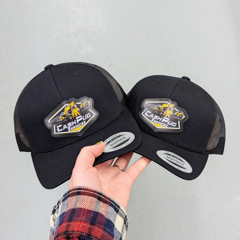 May include: Two black snapback trucker hats with mesh backs. Each cap features a hexagonal patch with a yellow cartoon pug character and the text "CASH PUG SERVICES" in white and yellow. Both hats have a silver "SNAPBACK" sticker on the brim.
