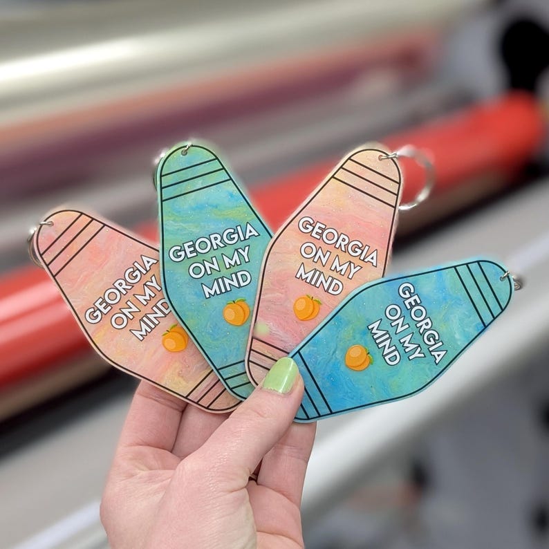 May include: Four motel-style keychains featuring a marbled finish in shades of pink and blue. Each keychain displays the text "GEORGIA ON MY MIND" in white capital letters, accompanied by a small orange peach graphic near the bottom edge.