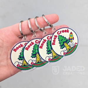 May include: Four circular clear acrylic keychains featuring a graphic of a green pine tree and a yellow rocket ship with the text Rock Creek Elementary in red letters. Each keychain has a silver metal split ring attached to the top for keys.