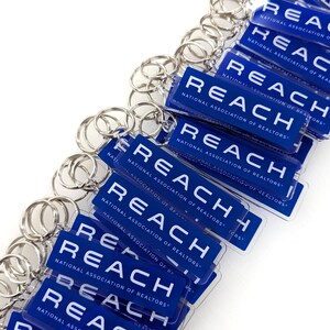 May include: A collection of rectangular blue keychains with silver metal rings. Each keychain features the white text REACH and the smaller text NATIONAL ASSOCIATION OF REALTORS printed on the front surface in a clean, sans-serif font style.