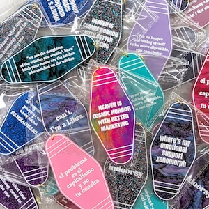 May include: A collection of motel-style keychains in various colors and finishes, including glitter, marble, and solid plastic. Each keychain features white printed text with phrases like 'Heaven is cosmic horror' and 'Where's my emotional support xenomorph'.