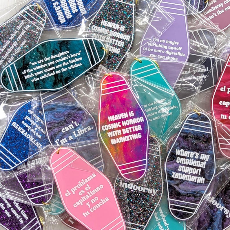 May include: A collection of motel-style keychains in various colors and finishes, including glitter, marble, and solid plastic. Each keychain features white printed text with phrases like 'Heaven is cosmic horror' and 'Where's my emotional support xenomorph'.