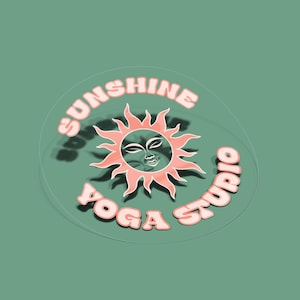 May include: A circular transparent sticker featuring a stylised sun graphic with a human face at the centre. The text SUNSHINE YOGA STUDIO is printed in a curved, bold, light pink font around the sun icon, which also has pink rays.