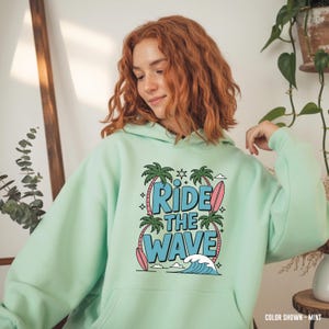 May include: A mint green hoodie featuring the text "Ride the Wave" in a graphic design with palm trees, a surfboard, and a wave. The hoodie has a kangaroo pocket and a drawstring hood.