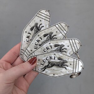 Op de afbeelding: Vier witte parelmoerkleurige sleutelhangers in motelstijl met een zwarte inktillustratie van een rennend konijn en het woord LUCKY in het zwart gedrukt. Elke sleutelhanger heeft dunne zwarte lijnen en kleine steraccenten.