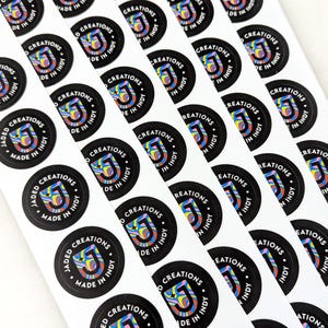 May include: A sheet of circular stickers featuring a black background with a central letter J filled with a multicolored swirling pattern. The text JADED CREATIONS MADE IN INDY is printed in white capital letters around the circular border.