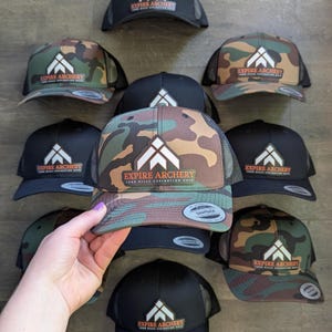 May include: A collection of snapback hats in solid black and camouflage patterns. Each hat features a central patch with a white geometric arrow logo and the text EXPIRE ARCHERY YOUR KILLS EXPIRATION DATE in orange and white lettering.
