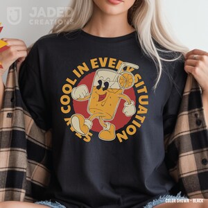 May include: Black t-shirt featuring a vintage cartoon of a juice drink with a straw and orange slice. The text around the image reads "Stay Cool in Every Situation".