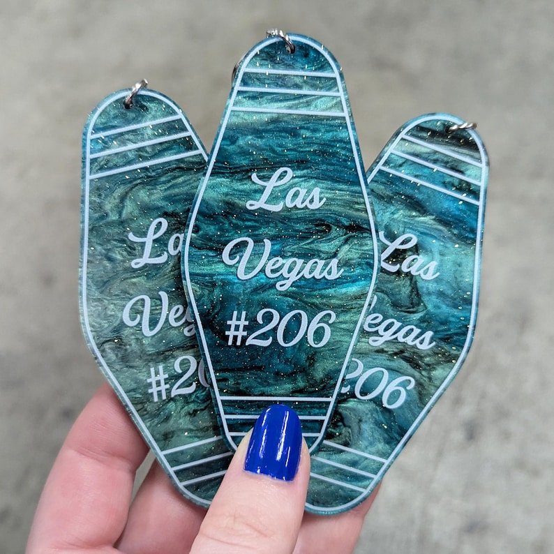 May include: Three motel-style keychains made of teal and black marbled acrylic with glitter accents. Each keychain features white text reading Las Vegas #206, horizontal white lines, and a small metal jump ring at the top for attachment.