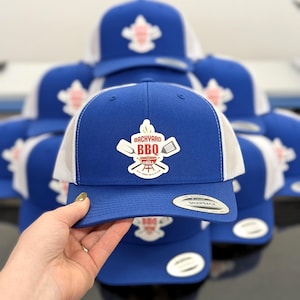 Puede incluir: Una gorra tipo camionero de color azul real y blanco con parte trasera de malla y un parche frontal con el texto BACKYARD BBQ GRILL, acompañado de ilustraciones de una espátula, un volteador y una parrilla. La visera incluye una pegatina plateada.