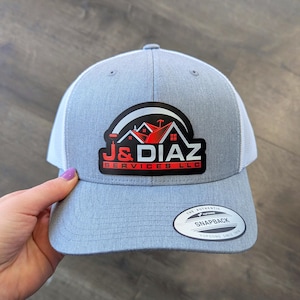 May include: A heather gray and white trucker-style baseball cap featuring a black patch on the front with a red and white logo that reads J&DIAZ SERVICES LLC. The brim includes a circular sticker with the text THE AUTHENTIC YUPOONG SNAPBACK.