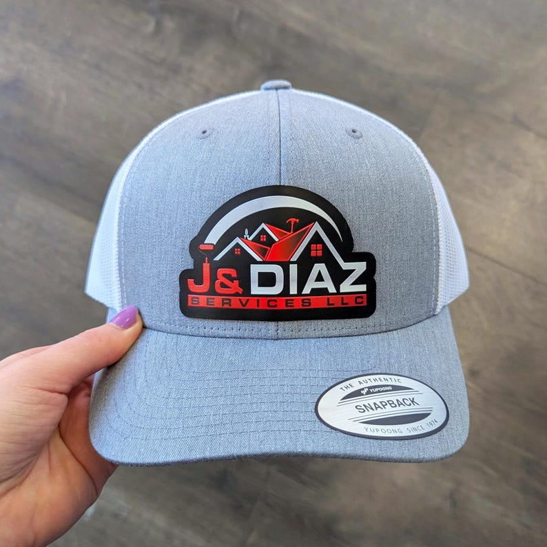 May include: A heather gray and white trucker-style baseball cap featuring a black patch on the front with a red and white logo that reads J&DIAZ SERVICES LLC. The brim includes a circular sticker with the text THE AUTHENTIC YUPOONG SNAPBACK.
