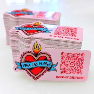 May include: A stack of rectangular pink stickers featuring a red heart with a yellow flame, a blue banner with the text VIVA LAS FLORES, a red QR code, an Instagram icon, and the red text @VIVALASFLORESFLORIST printed on the right side of each sticker.