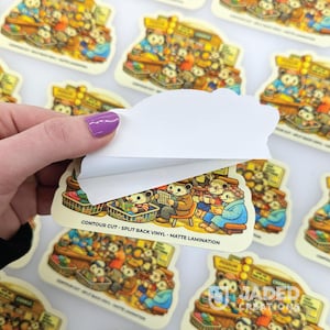 May include: A die-cut vinyl sticker featuring a colorful illustration of anthropomorphic animals in a market setting. The sticker has a matte finish and a split-back paper backing partially peeled away, revealing the text CONTOUR CUT - SPLIT BACK VINYL - MATTE LAMINATION.