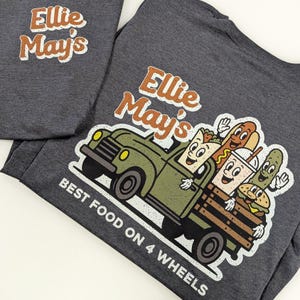 May include: Two dark grey t-shirts. One displays the text "Ellie May's" in orange and white. The other features a cartoon truck loaded with food, including a hot dog and burger, with the text "Best Food on 4 Wheels".