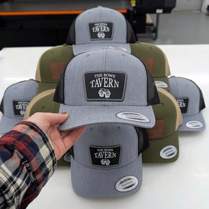 May include: A collection of snapback hats featuring a heather gray front panel and black mesh back. Each hat displays a black rectangular patch with the white text THE BOWE TAVERN and a graphic of two clinking beer mugs centered on the front.