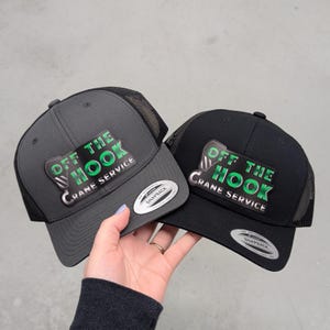 May include: Two snapback trucker hats, one dark gray and one black, featuring mesh back panels and a front patch with a crane hook graphic and the text "OFF THE HOOK CRANE SERVICE" in green and silver. Each hat has a round silver sticker on the brim.